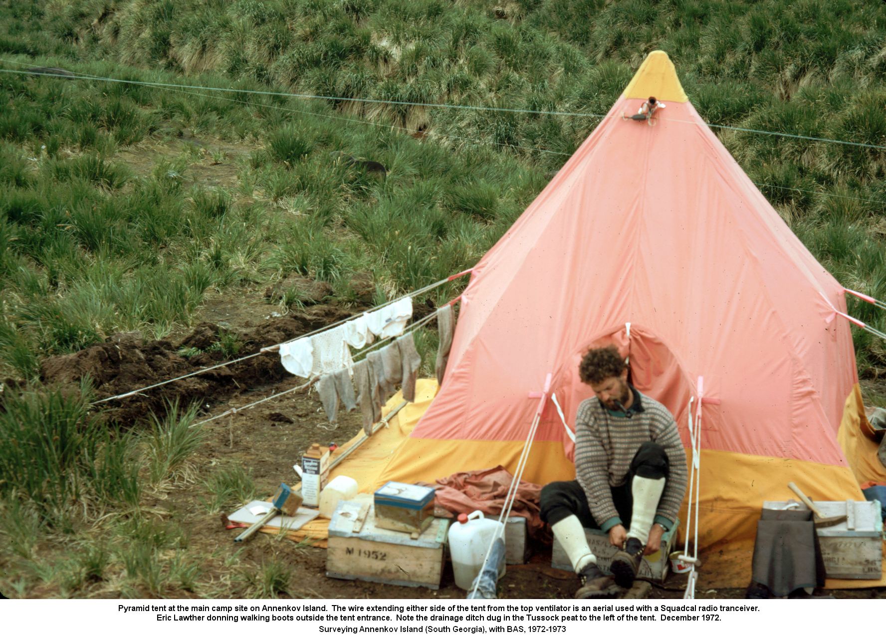 Pyramid tent at the main camp site on Annenkov Island.  The wire extending either side of the tent from the top ventilator is an aerial used with a Squadcal radio tranceiver. Eric Lawther donning walking boots outside the tent entrance.  Note the drainage ditch dug in the Tussock peat to the left of the tent, December 1972.