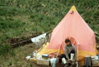 Pyramid tent at the main camp site on Annenkov Island. The wire extending either side of the tent from the top ventilator is an aerial used with a Squadcal radio tranceiver. Eric Lawther donning walking boots outside the tent entrance. Note the drainage ditch dug in the Tussock peat to the left of the tent, December 1972.